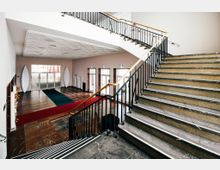 Geräumiges Treppenhaus mit Marmorboden und Treppe, die seitlich nach oben führt, geländer mit schwarzem Metallgeländer und Holzhandlauf. Im Hintergrund eine Glas-Doppeltür mit großen, verzierten seitlichen Elementen und rotem sowie blauem Teppich im Eingangsbereich.