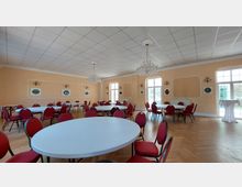 Ein festlich eingerichteter Saal mit runden und rechteckigen Tischen, die mit roten gepolsterten Stühlen umgeben sind. Der Raum hat helle Wände, große Fenster mit Glasflächen, zwei hängende Kronleuchter und dekorative Wandverzierungen.