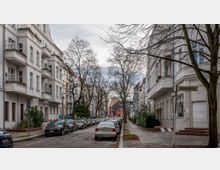 Straßenszene in einem Wohngebiet mit mehrstöckigen Altbauten aus der Gründerzeit, teils mit Balkonen und dekorativen Fassaden. Entlang der Straße sind parkende Autos und kahle Bäume; ein Straßenschild trägt die Aufschrift „Wolfsburger Straße“.