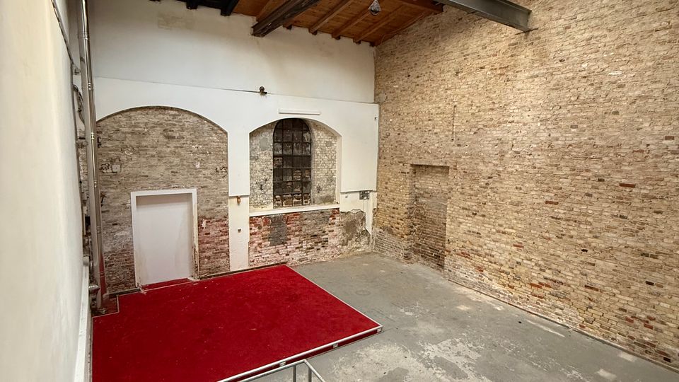 Innenraum einer ehemaligen Industriehalle mit hohen Ziegelwänden, sichtbaren Stahlträgern und großem Oberlicht im Dach. Unten ein Betonboden mit roter Plattform sowie eine Metalltreppe und ein weißer Wandbereich mit Rundbögen und einem vergitterten Fenster.