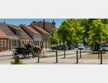 Straßenszene in einer Kleinstadt mit niedrigen, pastellfarbenen Häusern und roten Ziegeldächern entlang der Fahrbahn. Rechts säumen große, dicht belaubte Bäume den Straßenrand mit geparkten Autos, Pollern und einem Gehweg; im Hintergrund ragt ein Kirchturm über die Dächer.