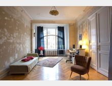 Alt-Text: Helles Wohnzimmer mit abgeblätterten Wänden, einem großen Bogenfenster mit dunklen Vorhängen, Holzdielenboden und mehreren Möbelstücken, darunter ein Sofa, ein Sessel, ein Schreibtisch mit Computer und ein kleiner Beistelltisch mit einer Vase voller roter Blumen.