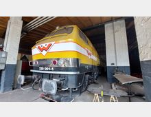 Gelbe Diesellokomotive mit weißen und roten Streifen, abgestellt in einem überdachten Lokschuppen mit weiß gestrichenen Backsteinwänden und Holzdach. Vor der Lok befinden sich Werkzeuge und Bauteile.