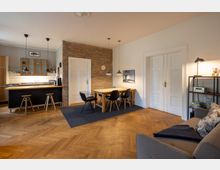 Offener Wohn- und Essbereich mit hellem Fischgrätparkett, einer Küche mit Theke und Holzfronten, einem Esstisch aus Holz mit schwarzen Stühlen sowie weißen Türen und einer Ziegelsteinwand. Beleuchtung erfolgt durch mehrere Hängeleuchten über Esstisch und Küchenbereich.