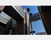 Moderne Bürogebäude mit großen Glasfassaden und verbindenden Brücken, die über die Gebäude führen, im Blick von unten; eine Straßenlaterne und ein kleiner Teil eines weiteren Gebäudes mit „Scandic“-Schriftzug im Hintergrund sichtbar.