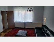 Wohnzimmer mit weiß gestrichenen Wänden, einem großen Fenster mit weißen Vorhängen, einer schwarzen Ledersofa, einem roten Teppich und einem kleinen schwarzen Hocker. An der Decke hängt eine verzierte Lampe, und unter dem Fenster befindet sich ein Heizkörper.