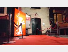 Eingang eines Kinos mit rotem Teppich, umgeben von Absperrungen mit Samtbändern. Im Hintergrund sind helle Klinkerwände, eine schwarze Tür sowie ein beleuchtetes rotes Schild mit dem weißen Logo der Berlinale und der Schrift „Berlinale“ zu sehen.