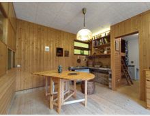 Holzverkleideter Raum mit einem ovalen Holztisch in der Mitte. An der Wand befinden sich Regale mit Büchern und Dekorationsgegenständen, ein kleines Fenster mit gelbem und blauem Glas sowie eine geöffnete Tür, die in einen angrenzenden Raum mit einer Leiter führt.