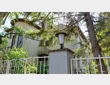 Zweistöckiges Wohngebäude mit hellgelber Fassade, hohen Fensterfronten und einem Satteldach, umgeben von Bäumen und dichtem Grün. Im Vordergrund ein Zaun mit einer Säule, auf der eine Außenlaterne installiert ist.