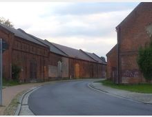 Straßenszene mit alten, langgezogenen Backsteingebäuden auf beiden Seiten einer leicht geschwungenen, asphaltierten Straße. Die Gebäude haben hohe, teils zugemauerte Rundbogentüren und sind mit Dachziegeln bedeckt, teilweise mit Graffiti und Efeu bewachsen.