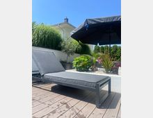 Sonnenliege auf einer Holzterrasse, umgeben von grünen Pflanzen in Blumenbeeten mit weißen Steinen. Im Hintergrund ein schwarzer Sonnenschirm und ein modernes Wohnhaus mit weißer Fassade.