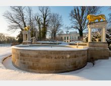 Ein verschneiter Garten mit einem Rundbrunnen aus Sandstein im Vordergrund, flankiert von zwei Säulen mit goldenen Löwenfiguren darauf. Im Hintergrund befindet sich ein klassizistisches Gebäude mit großen Fenstern und grünen Fensterläden, umgeben von hohen Bäumen.