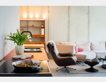 Offener Wohn- und Küchenbereich mit Betonwänden, einer Kücheninsel mit Kochfeld und Pfanne sowie einem Regal mit Beleuchtung im Hintergrund. Daneben stehen ein Ecksofa, ein Leder-Loungesessel mit Hocker und ein niedriger Couchtisch auf einem Teppich.