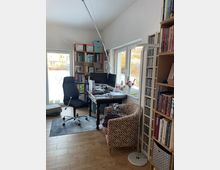 Helles Arbeitszimmer mit Holzfußboden, Schreibtisch mit zwei Monitoren und Schreibtischlampe sowie einem Drehstuhl auf einem Teppich. An den Wänden stehen hohe Regale mit Büchern und CDs; große Fenster und eine Glastür öffnen den Blick in einen Garten.