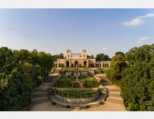 Historisches Gebäude mit symmetrischer Architektur, zwei Türmen und Arkaden, umgeben von gepflegten Gärten und Treppenanlagen; eingebettet in eine grüne Parklandschaft unter klarem Himmel.