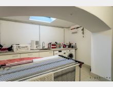 Waschküche mit weiß gefliesten Wänden und Boden, ausgestattet mit einer Waschmaschine, einem Waschbecken und einer Arbeitsfläche mit Reinigungsmitteln. Ein Wäschekorb, Wäscheständer und ein Oberlicht sind ebenfalls sichtbar.