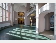 Innenraum eines historischen Gebäudes mit hohen Decken, großen, bogenförmigen Fenstern und geschwungenen weißen Geländern im Jugendstil. Das grünliche Parkett und die Lichtreflexionen verleihen der Halle eine helle, offene Atmosphäre.