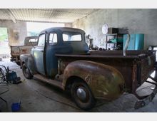 Eine Werkstatt mit einem alten, rostigen Pickup-Truck im Vordergrund. Im Hintergrund stehen weitere Werkzeuge, Geräte und Fahrzeugteile, sowie ein zweiter, ebenfalls gealterter Truck.