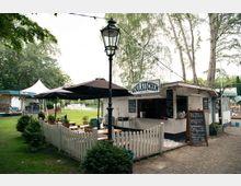 Ein kleiner Imbissstand mit der Aufschrift „Soulkitchen“ steht in einer grünen Parkumgebung, umgeben von Bäumen. Der Bereich ist mit weißen Holzzäunen, Tischen, Sitzplätzen unter Sonnenschirmen und einer Schiefertafel mit Speiseangebot gestaltet.