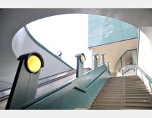 Eine Treppe mit seitlich angebrachtem Geländer und runden Leuchten führt zu einem modernen Gebäude mit gläserner Fassade. Daneben verläuft eine Rolltreppe, und im Hintergrund ist ein Teil eines großen Uhrendesigns sichtbar.