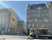 Straßenszene mit historischen Gebäuden, darunter ein Sandsteinbau mit Gesimsen und Türmchen sowie ein Nachbargebäude mit moderner Verglasung. Im Vordergrund befinden sich Ampeln, eine leere Straßenkreuzung, ein Lastwagen und ein Roller mit zwei Personen.