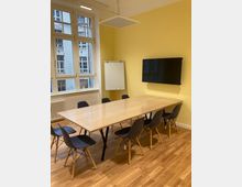 Konferenzraum mit einem langen, hellen Holztisch und acht schwarzen Stühlen mit Holzbeinen auf einem Holzboden. An der gelben Wand befinden sich ein Flachbildschirm und ein weißes Flipchart, daneben große Fenster mit Blick auf eine benachbarte Ziegelhausfassade.