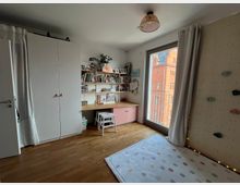 Ein Kinderzimmer mit Holzboden, einem weißen Kleiderschrank, einem Schreibtisch mit Regalen voller Bücher und Dekorationen sowie einem Fenster mit Blick auf ein rotes Backsteingebäude. An der rechten Wand befindet sich eine Kletterwand, und auf dem Boden liegt eine bunte Punktedecke.