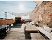 Holzterrasse mit gemütlichen Sitzmöbeln, einem Esstisch aus schwarzem Metall und Sonnensegeln, flankiert von einer hohen Ziegelwand und einem Bambuszaun; im Hintergrund steht eine kleine Holzhütte.