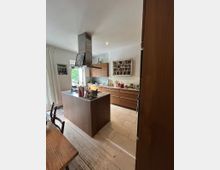 Moderne Küche mit Holzschränken und einer zentralen Kochinsel. Im Hintergrund befinden sich eine Gewürzablage an der Wand, ein großes Fenster und helle Bodenfliesen; ein Esstisch mit Stühlen ist im Vordergrund sichtbar.