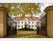 Eine historische Villa mit gelber Fassade und rotem Dach, umgeben von herbstlicher Landschaft. Im Vordergrund stehen geöffnete schmiedeeiserne Tore, darunter liegt ein mit gelben Blättern bedeckter Weg.