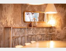 Gemütlicher Innenraum mit verputzten, ungleichmäßigen Wänden, einer schmalen Holzablage und hohen weißen Barhockern. Ein rechteckiger Spiegel mit goldenem Rahmen und Dekoration mit getrockneten Blumen in Vasen stehen im Vordergrund, beleuchtet von einer Tisch- und Pendelleuchte.