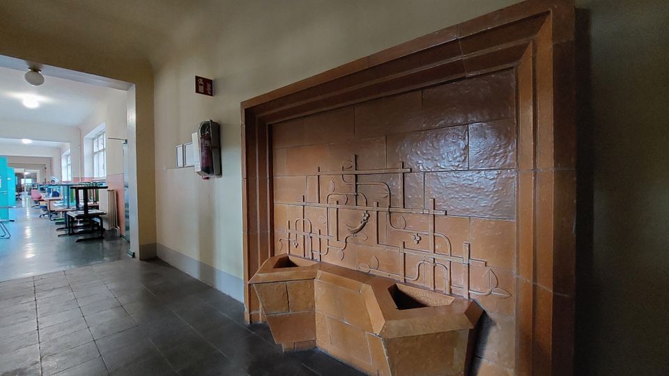 Ein Innenraum mit einem braunen Wandbrunnen aus Keramik mit geometrischen Mustern. Im Hintergrund ein Flur mit Tischen, Stühlen und blauen Trennwänden sowie Fenstern, die Tageslicht einlassen.