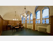 Historischer Raum mit gewölbter Decke, Holztäfelung an den Wänden und großen Fenstern mit Gitterstruktur. In der Mitte befindet sich ein Tisch mit Stühlen, und mehrere hängende Leuchter beleuchten den Raum.