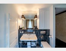 Modernes Badezimmer mit einem Waschbecken auf einem Metallgestell, dunkler Marmorverkleidung, großem Spiegel und zwei schwarzen Wandlampen. Rechts ein weißer Wandschrank, links ein Handtuchheizkörper und im Hintergrund ein Fenster mit beigefarbenen Vorhängen.