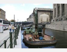 Straßenszene an einem Kanal mit einem schwimmenden Lastkran auf einem Ponton. Im Hintergrund befinden sich historische Gebäude aus massivem Stein, eine Brücke sowie geparkte Reisebusse, während Passanten auf einem Gehweg entlang der Uferpromenade spazieren.