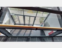 Blick auf den Eingangsbereich eines modernen Gebäudes mit Glasdoppeltüren, schwarzen Fliesen und einem Heizkörper an der weißen Wand. Draußen ist ein gepflasterter Weg sowie ein Grünstreifen sichtbar.