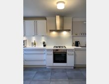 Helle, moderne Küche mit weißen Schränken und Schubladen, einer Edelstahl-Dunstabzugshaube über einem Gasherd mit Backofen. Auf der Arbeitsfläche stehen ein Messerblock, eine Flasche sowie eine Kaffeemaschine, daneben ein Einbauspülbecken. Der Boden ist mit grauen Fliesen ausgelegt.