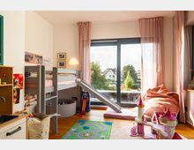 Kinderzimmer mit einem Hochbett aus Holz, einer Rutsche und hellrosa Betten. Vor einem großen Fenster mit rosa Vorhängen stehen ein Sitzsack, Spielzeug und eine Spielmatte mit Straßendesign; im Hintergrund ist eine grüne Außenansicht mit Häusern sichtbar.
