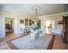 Ein eleganter Salon mit pastellfarbenen Wänden, verziert mit Wandgemälden und goldgerahmten Kunstwerken. Der Raum ist mit antiken Möbeln, einem Kronleuchter sowie einem floralen Teppich ausgestattet und führt durch geöffnete Türen in weitere Räume mit ähnlicher Einrichtung.