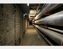 Industriegang mit einer schmalen Betonwand auf der linken Seite und mehreren großen silbernen Rohren, die an der rechten Wand befestigt sind. Die beleuchtete Decke zeigt Kabel und Rohrleitungen, und der Gang führt in einen dunkel wirkenden Bereich im Hintergrund.