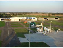 Kleiner Flugplatz mit mehreren geparkten Flugzeugen auf einer asphaltierten Fläche, einem großen Hangar mit blauer Beschriftung im Hintergrund und umgeben von Feldern und Wiesen. Im Vordergrund befinden sich weiße Treibstofftanks und gepflasterte Abstellflächen.