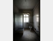 Ein verlassener, heruntergekommener Badezimmerraum mit gefliesten Wänden und Fußboden. Zu sehen sind eine Duschkabine mit halbtransparenten Wänden, eine Toilette vor einem Fenster mit beschädigtem Rahmen, sowie kaputte Rohre und abblätternde Farbe an den Wänden und der Decke.