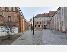 Historischer Innenhof mit Pflastersteinen, umgeben von Gebäuden aus Backstein und verputztem Mauerwerk, teilweise mit gotischen Merkmalen. Im Hintergrund sind moderne und historische Gebäude sowie ein kahler Baum im Vordergrund zu sehen.