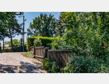 Ein gepflasterter Weg führt an einem Holzzaun vorbei, der von üppigem grünen Laub und blühenden Sträuchern umgeben ist; im Hintergrund stehen hohe Bäume und der Straßenschild „Neckarstraße“ ist sichtbar.