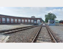 Ein Bahndepot mit einem halbkreisförmigen Lokschuppen aus rotem Backstein und geschlossenen Toren. Im Vordergrund verlaufen Schienen, die zu einer Drehscheibe führen; rechts steht ein kleines Kontrollhäuschen und abgestellte Züge sind sichtbar.