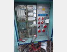 Ein geöffneter Verteilerkasten mit elektrischen Sicherungen, Schaltern und Steckdosen, darunter blaue und rote Industriesteckdosen. Mehrere Kabel und Stromanschlüsse führen aus dem Kasten, der sich in einer Außenumgebung befindet.