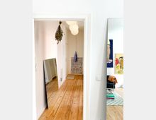 Blick durch eine Tür in einen hellen Flur mit Holzdielenboden, an dessen Ende ein kleines Regal mit CDs steht. Links lehnt ein großer Spiegel an der Wand, rechts ist der Teil eines Wohnzimmers mit bunten Kunstwerken und Möbeln sichtbar.