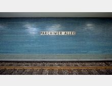 U-Bahn-Station mit Wandverkleidung aus blauen Fliesen und dem Stationsnamen „Parchimer Allee“ auf einem rechteckigen Schild in der Mitte; darunter Gleise mit Schotterbett.