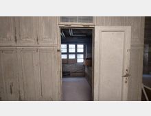 Alte Holztür mit abgeblätterter Farbe, die in einen kleinen, spärlich beleuchteten Raum mit mehreren Fensterscheiben führt; im Hintergrund sind eine Ziegelwand, eine Werkbank und Kartons zu sehen.