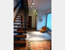 Moderne Wohnraumgestaltung mit einer offenen Holztreppe an der linken Seite, einem hellen Teppich auf dunklem Boden und dekorativen Orientteppichen. Im Raum befindet sich ein orangefarbener Sessel, eine Glasvase und eine Wanddekoration; darüber hinaus fällt Tageslicht durch ein großes Fenster auf der rechten Seite ein.